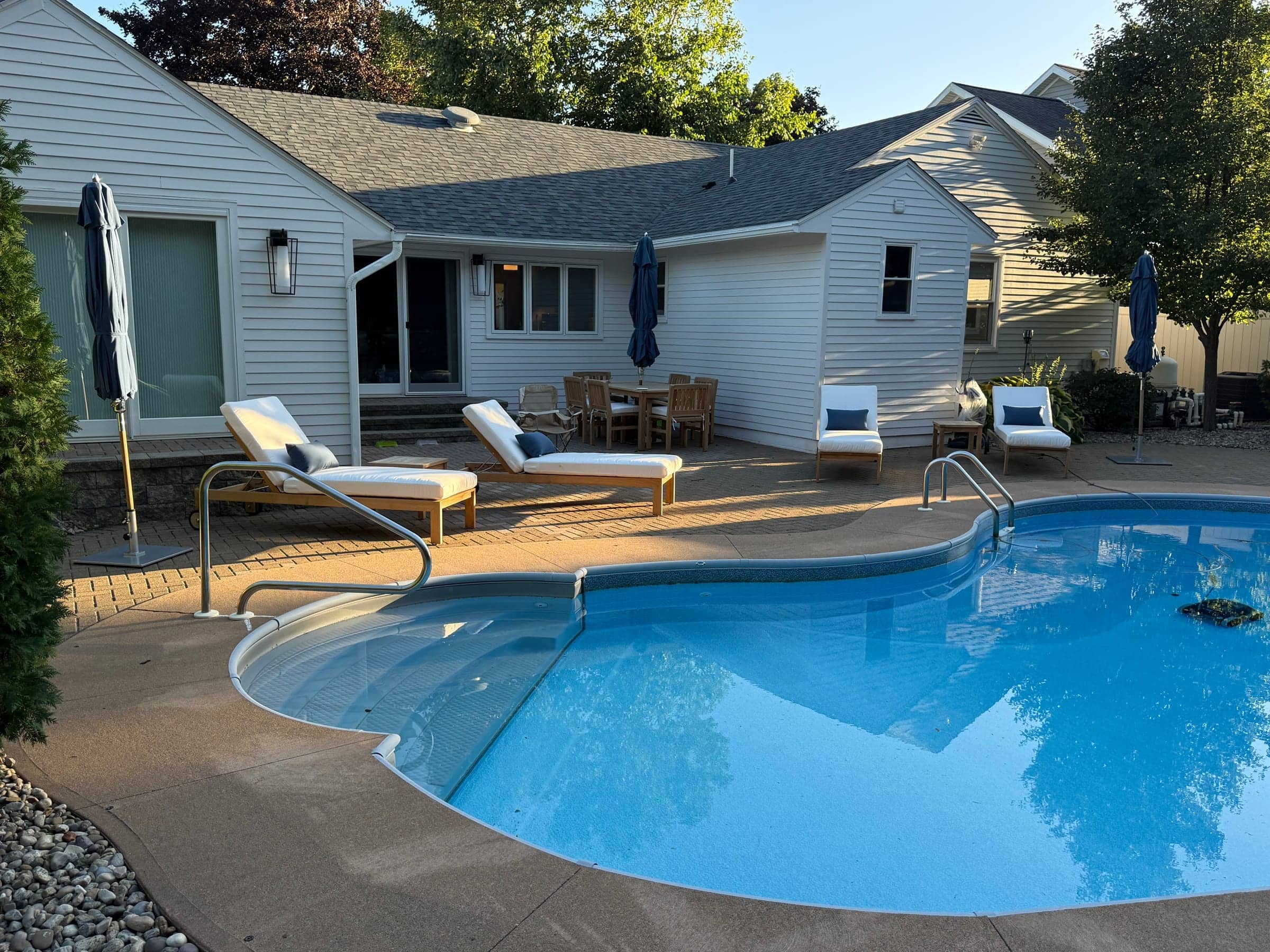 Wide view of a backyard pool with new lounge chairs, umbrellas and a teak dining set around the patio