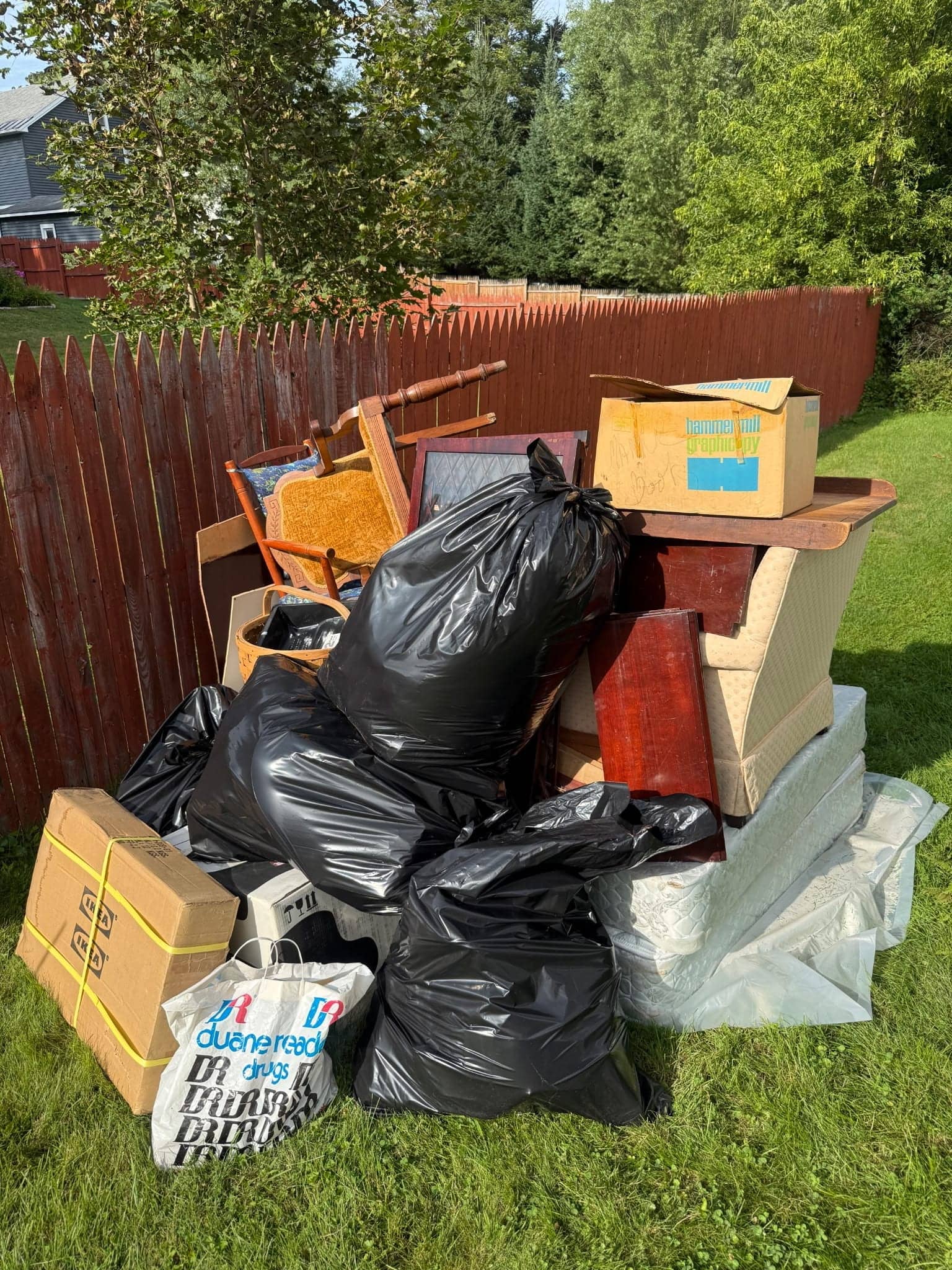 Pile of black trash bags, boxes, a mattress and old wooden furniture stacked against a fence in a yard