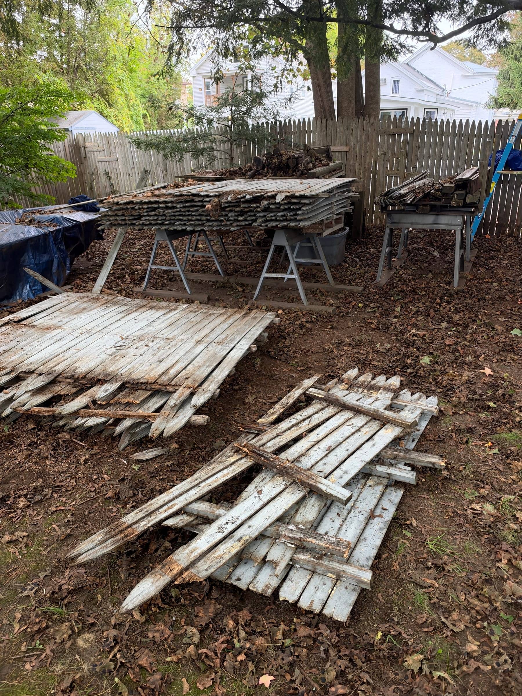 Old white picket fence panels torn out and stacked on the ground and on sawhorses in a fenced backyard