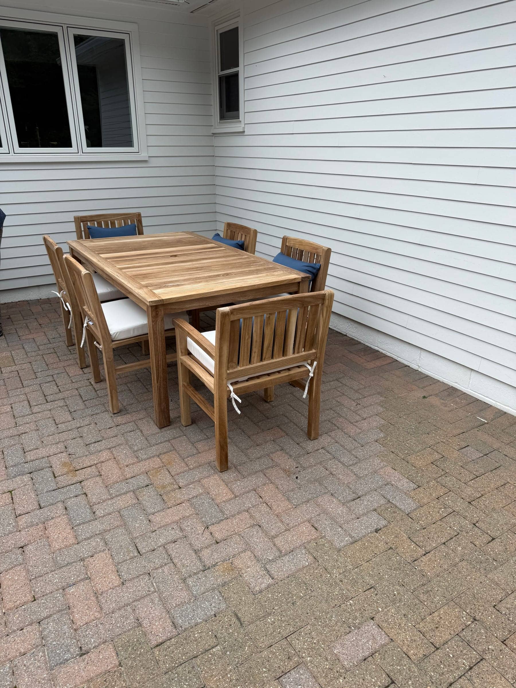 Teak outdoor dining table and six chairs set up on a brick patio beside a white house