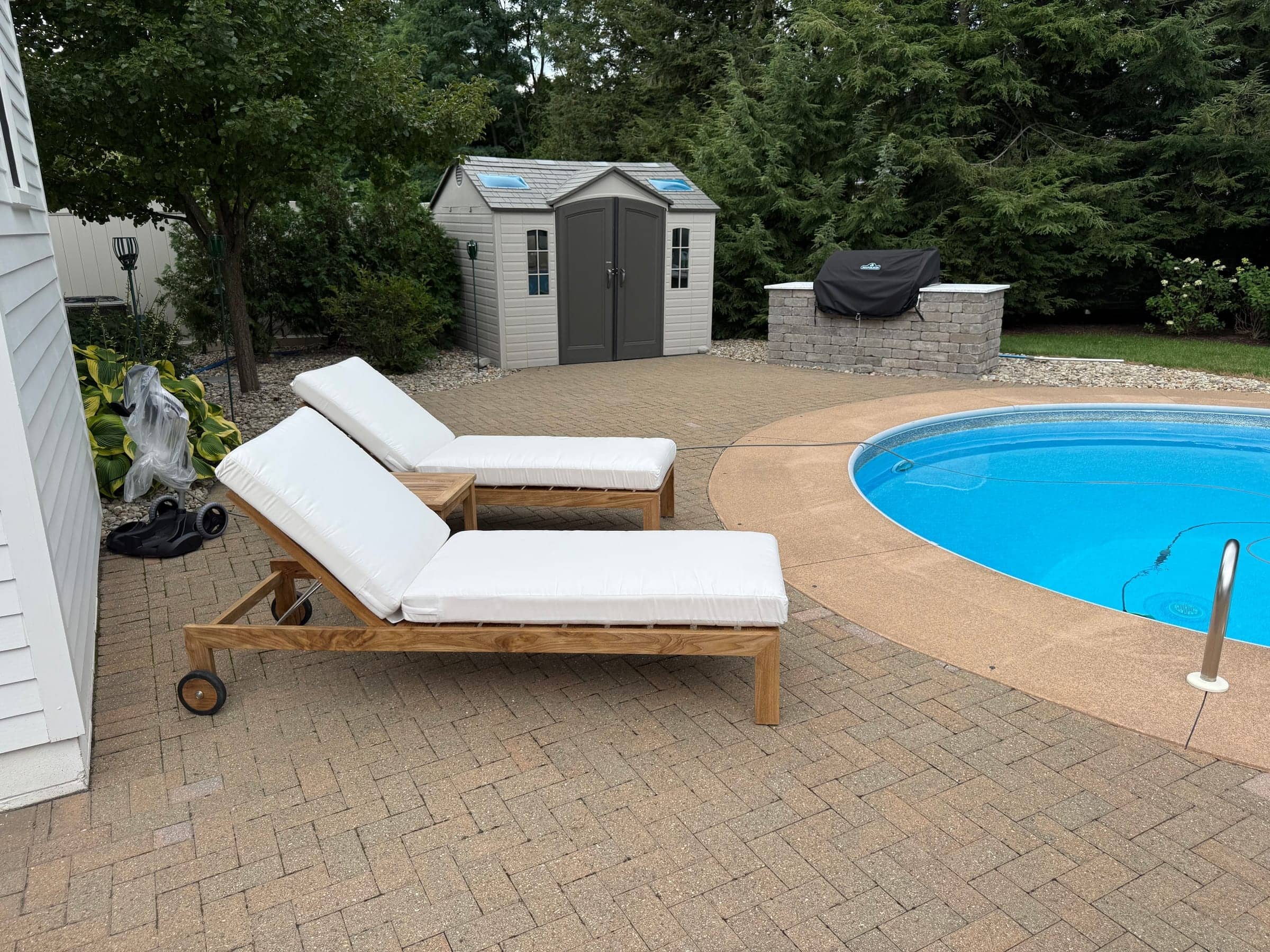 Two teak lounge chairs with white cushions set up on a brick patio beside an in-ground pool