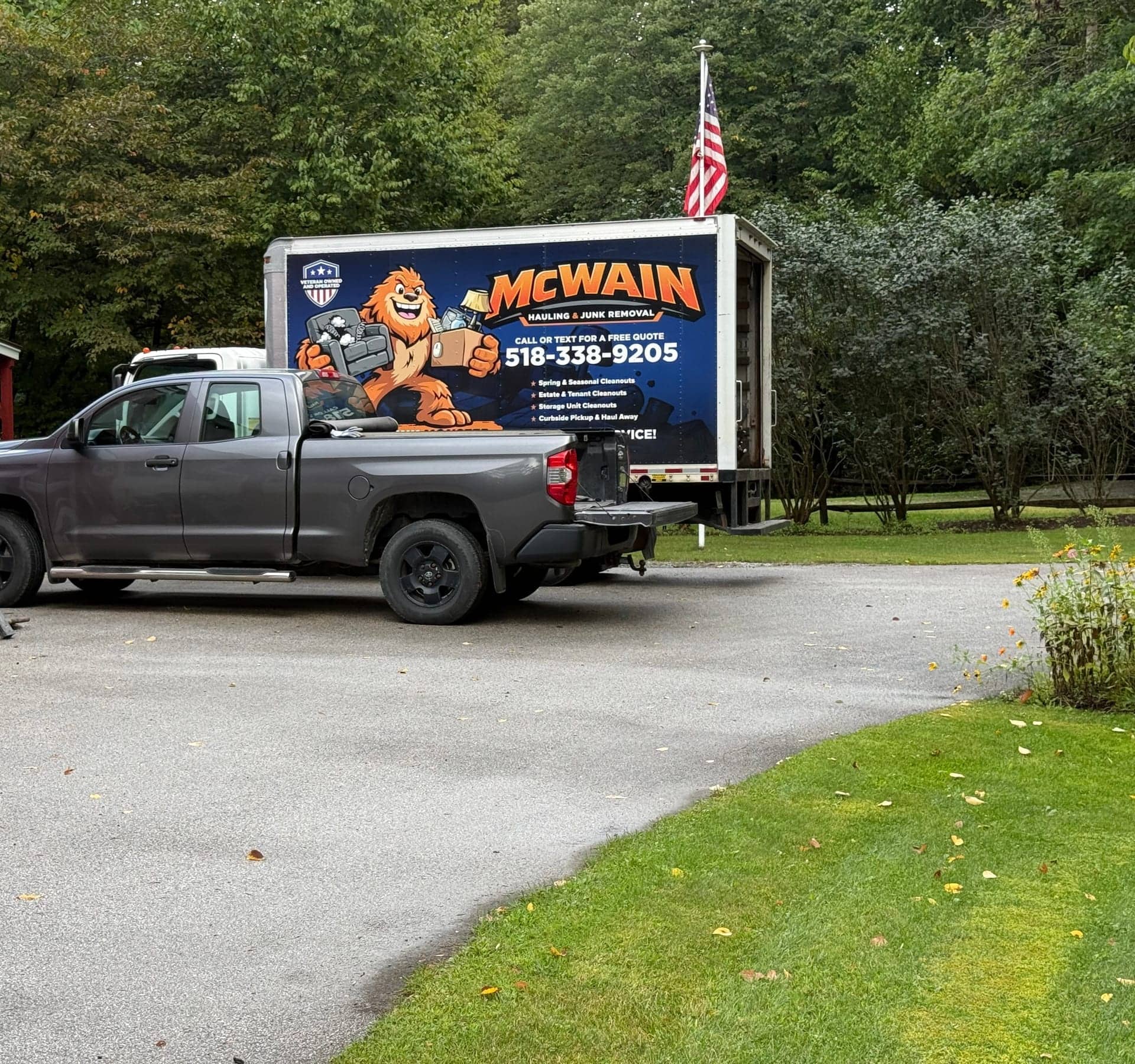 McWain Hauling box truck with the Junk Monster wrap parked behind a pickup in a paved driveway, American flag on a pole behind it